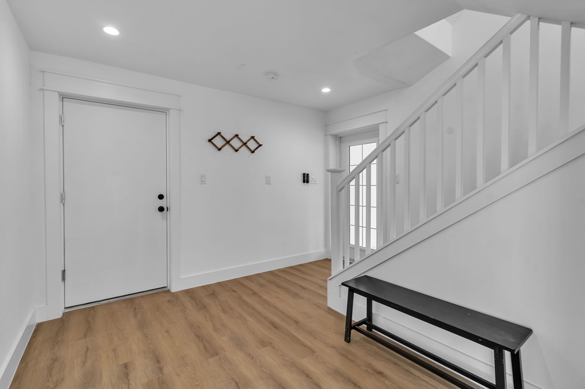A hallway with wooden flooring, a white door, a bench, and a staircase with white railings.