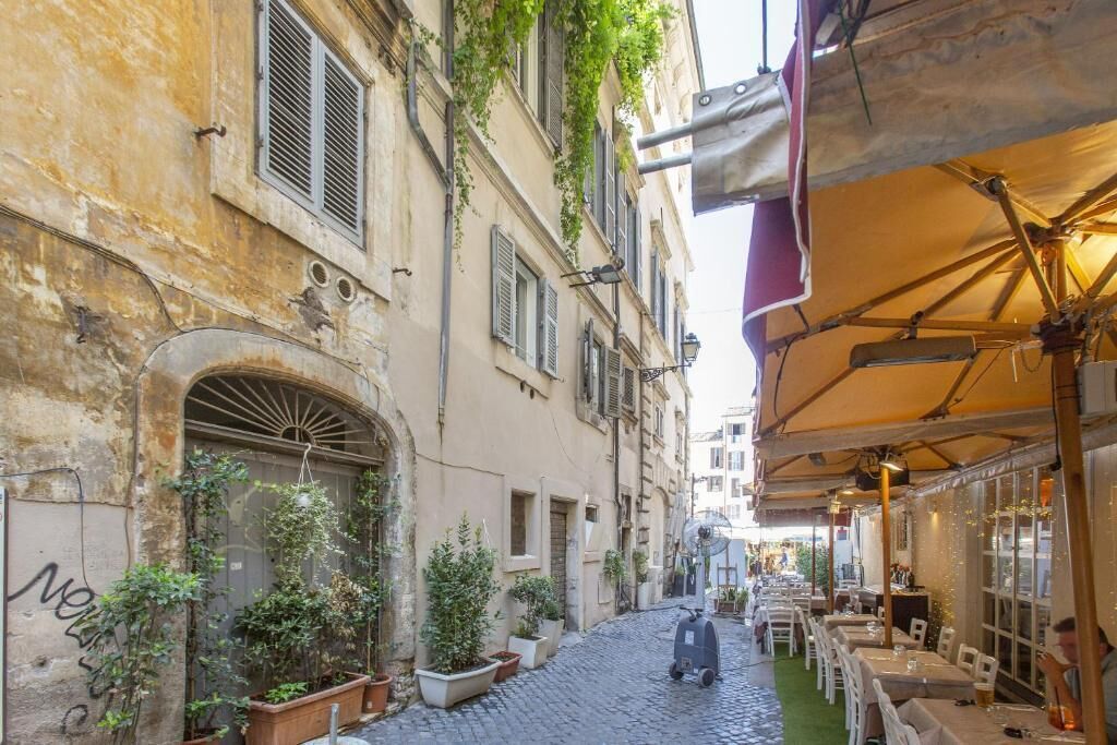 Una stretta strada acciottolata con tavolini all'aperto, piante in vaso e uno scooter parcheggiato a lato.