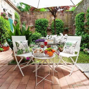 Une terrasse avec deux chaises blanches, une table garnie de fruits, de pâtisseries et d’une boisson, le tout entouré d’une végétation luxuriante et d’un mur de briques.