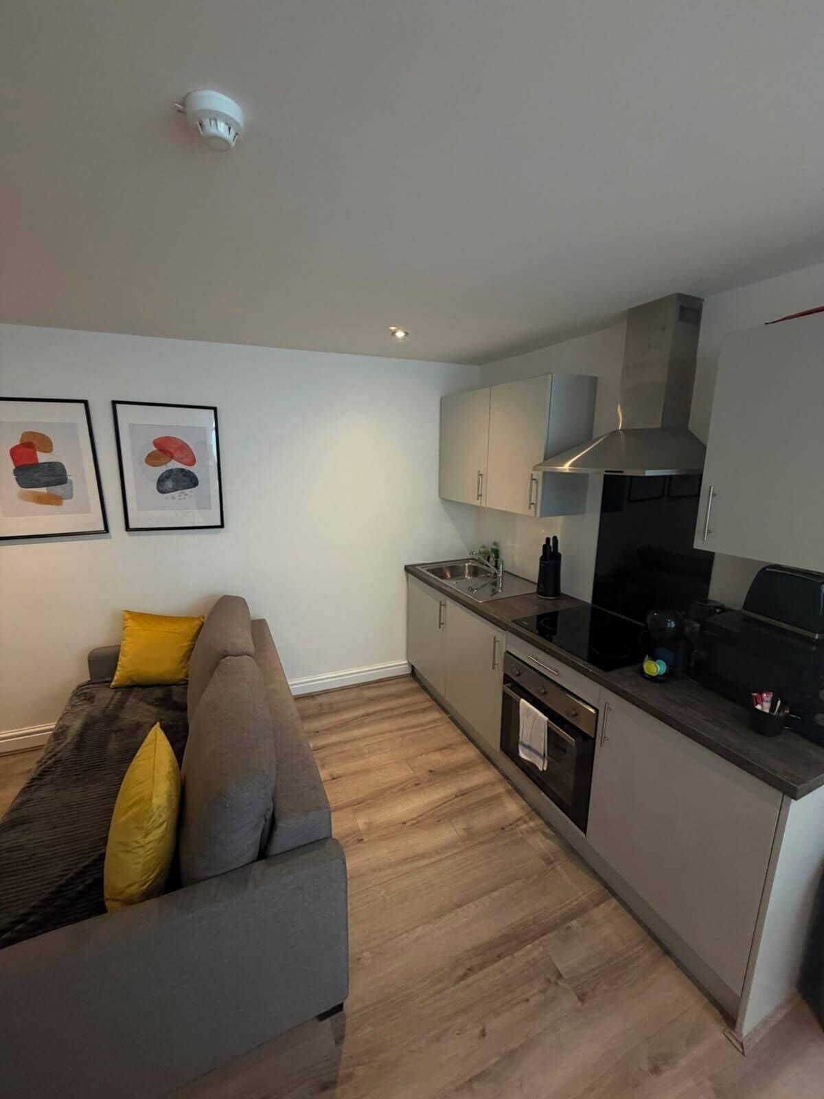 A modern kitchen with a built-in oven, a stovetop, and a range hood. There is a gray sofa with yellow cushions in the living area.