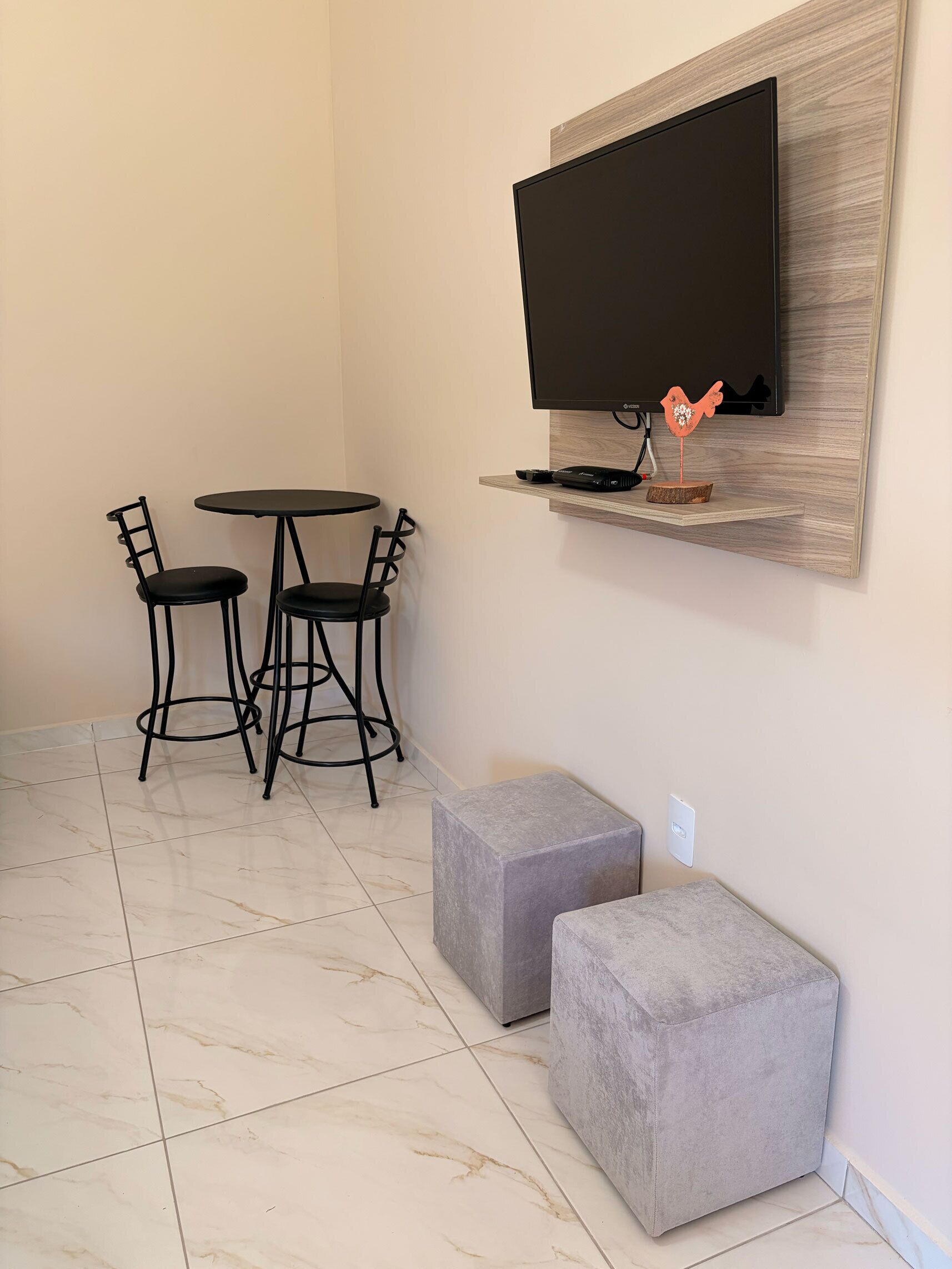 Quarto com televisão de tela plana fixada em uma prateleira de madeira, uma mesa redonda com duas cadeiras e dois pufes cinzas.