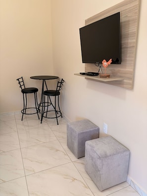 Una habitación con una televisión de pantalla plana montada en un estante de madera, una mesa redonda con dos sillas y dos otomanas grises.