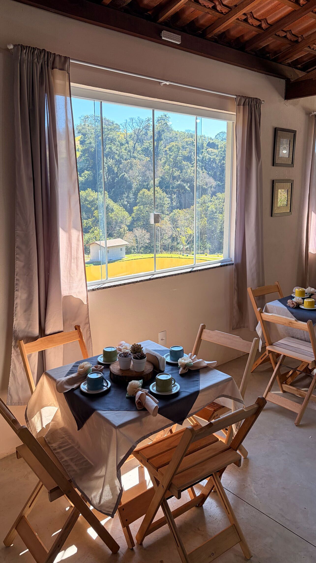 Un comedor con una mesa lista para dos personas, sillas de madera, vistas a un bosque a través de un ventanal grande y un techo con vigas a la vista.