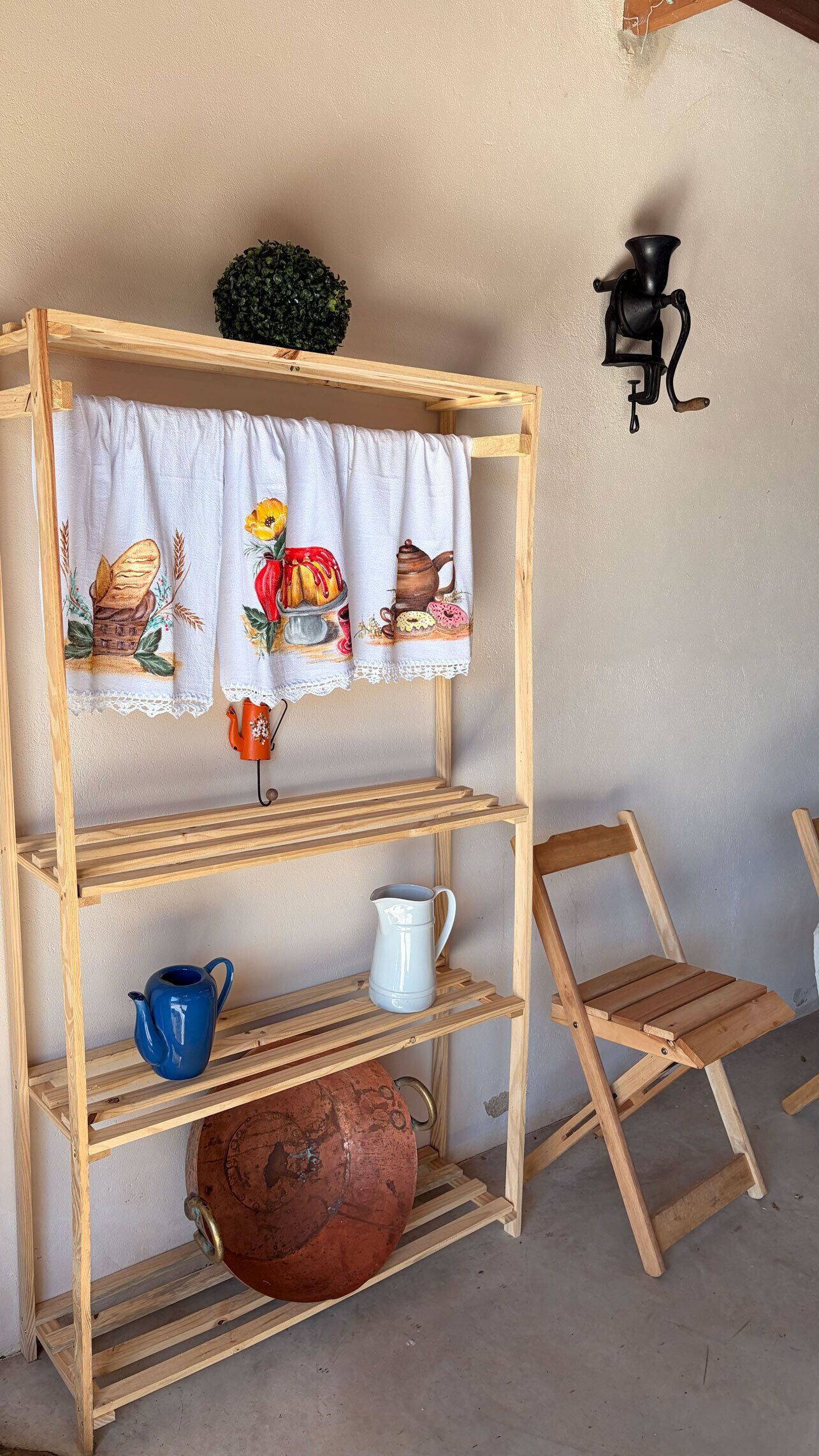 Una estantería de madera con un mantel blanco con ilustraciones de comida, una jarra azul, una jofaina blanca y una olla de cobre.