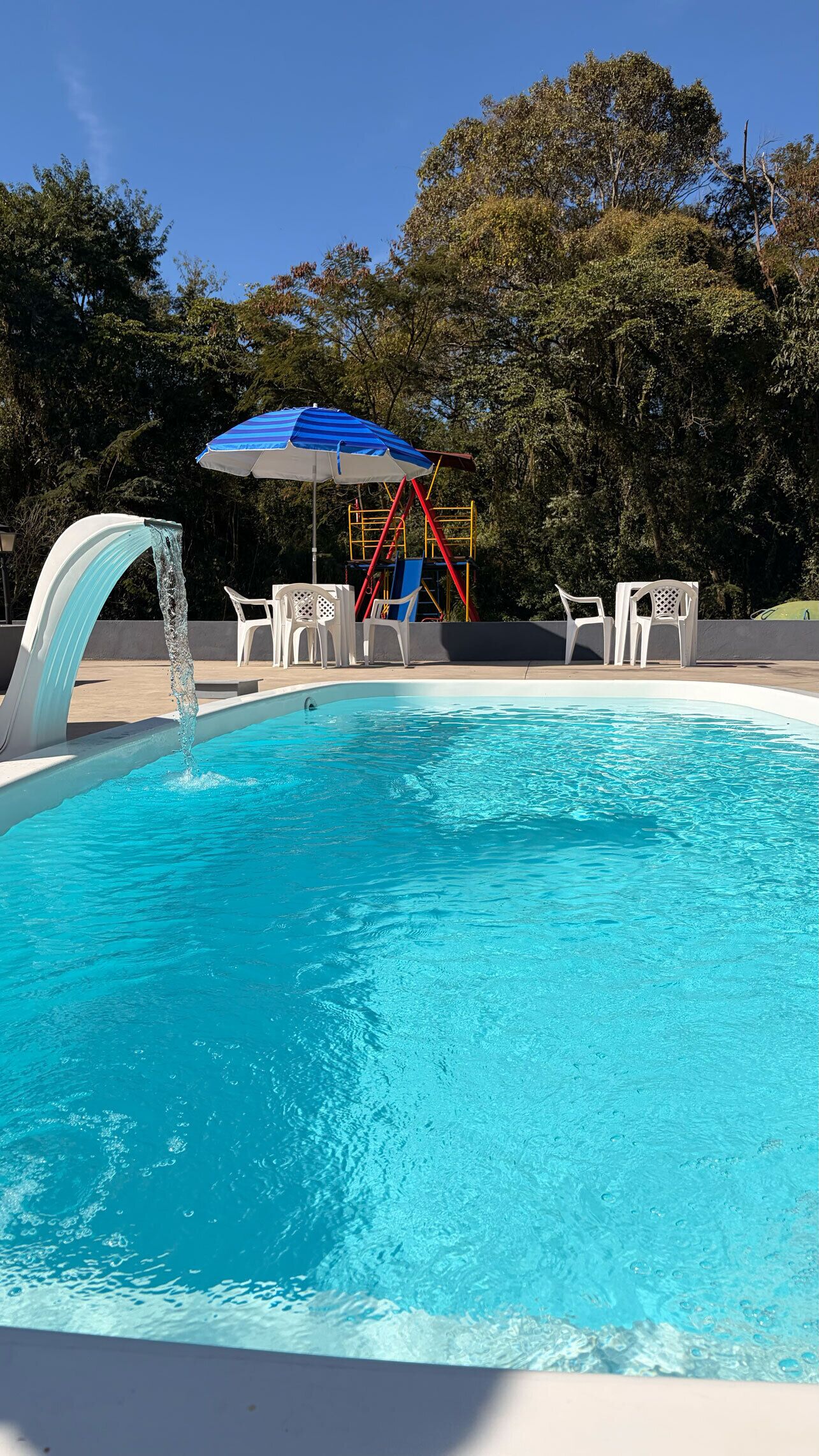 A swimming pool with a water feature, surrounded by white chairs and a playground structure.