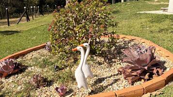 Un jardín con una estatua de cisne blanco, rodeado por un borde de ladrillo, con árboles y un cielo despejado al fondo.