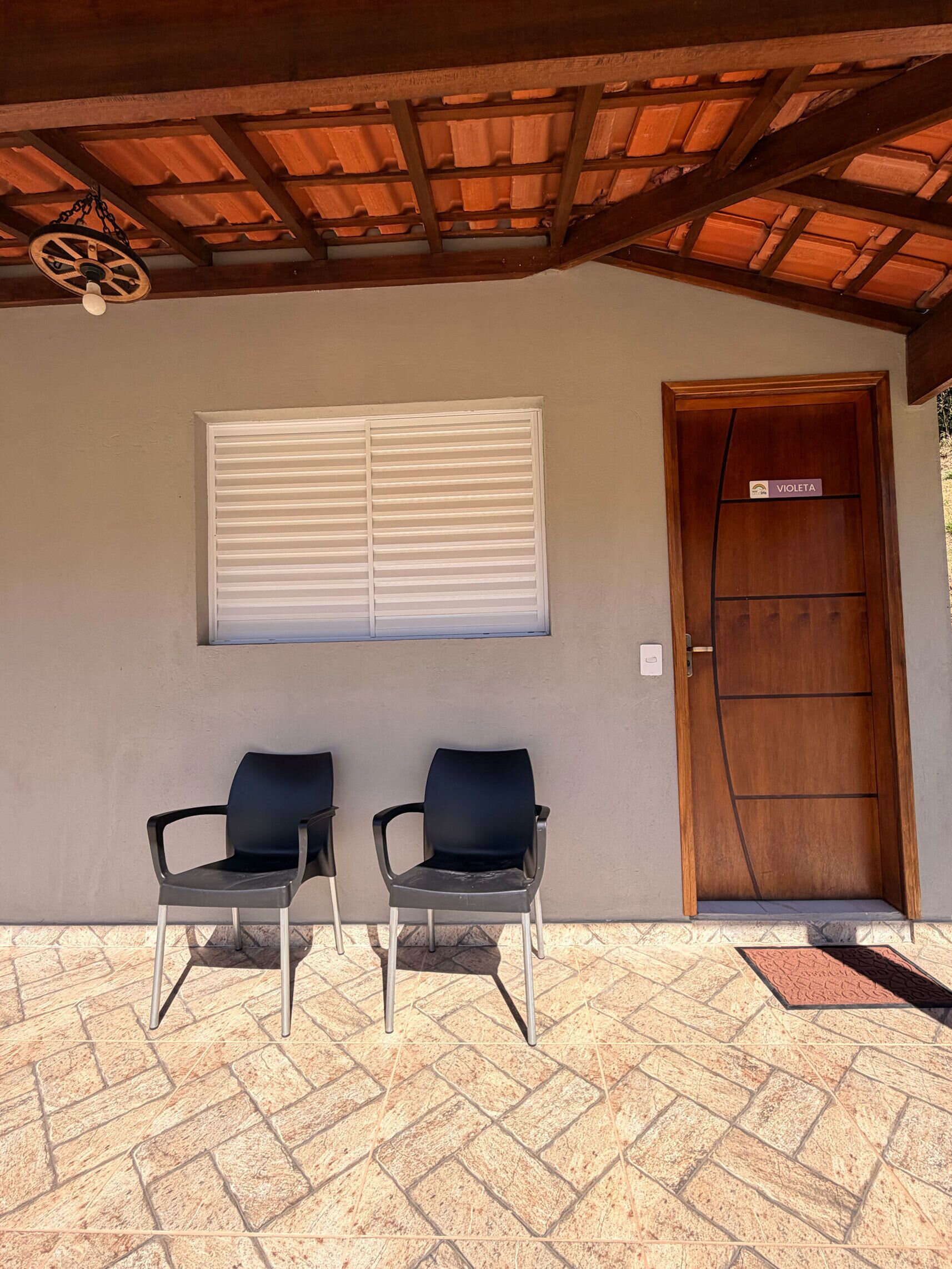 Un espacio de estar techado al aire libre con dos sillas, una ventana con persianas y una puerta de madera con una placa.