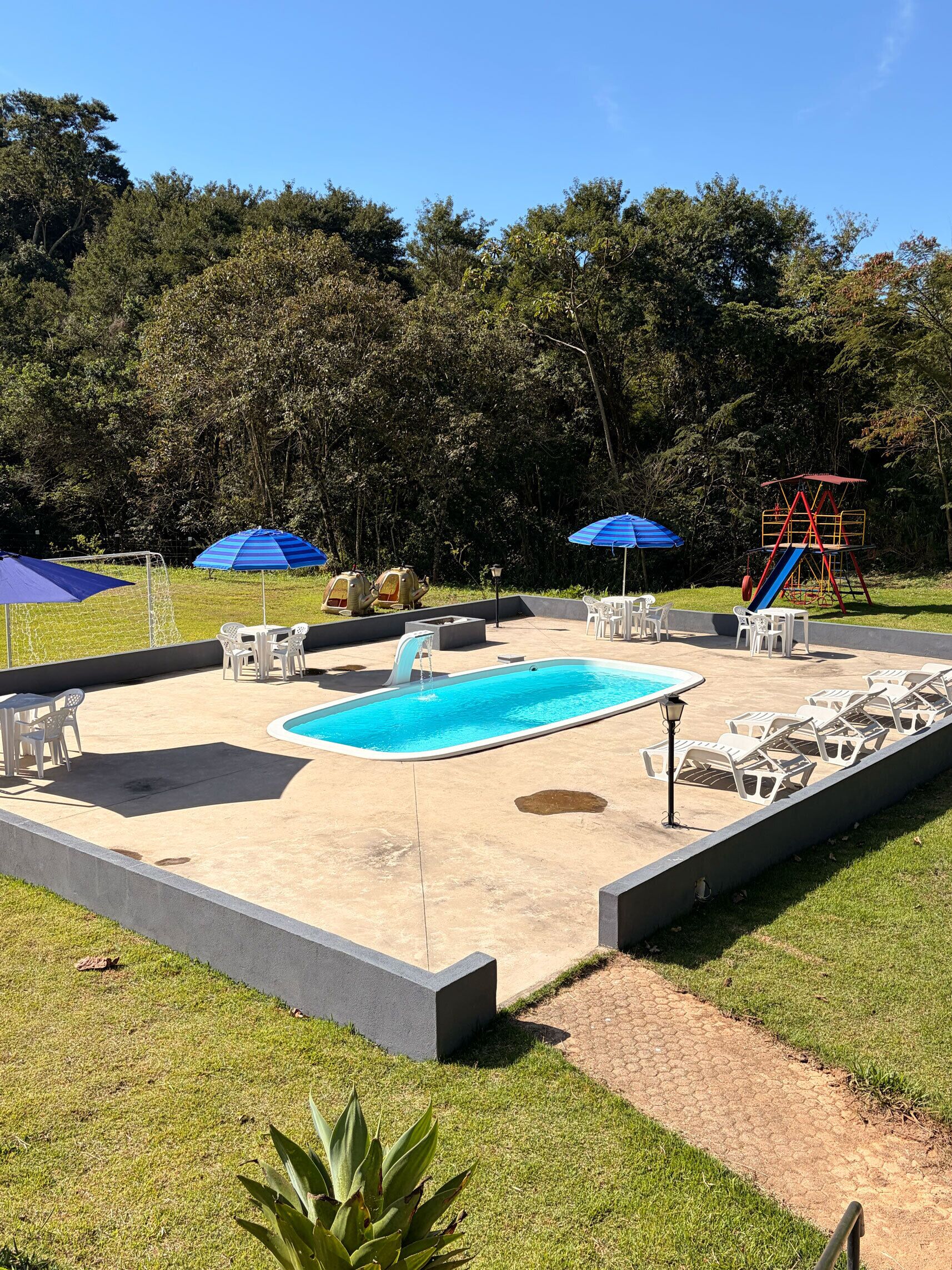 Una piscina con tobogán, rodeada de sillas de descanso y sombrillas blancas, con árboles al fondo.