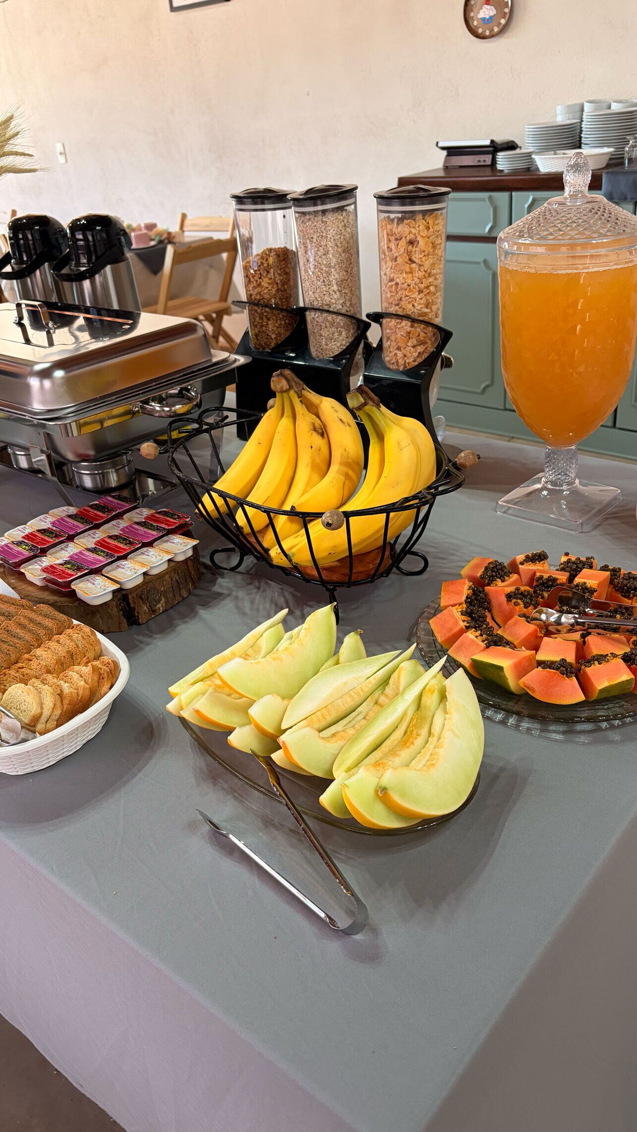 Una mesa de buffet con diversos alimentos, incluyendo plátanos, gajos de melón y un vaso de zumo de naranja.