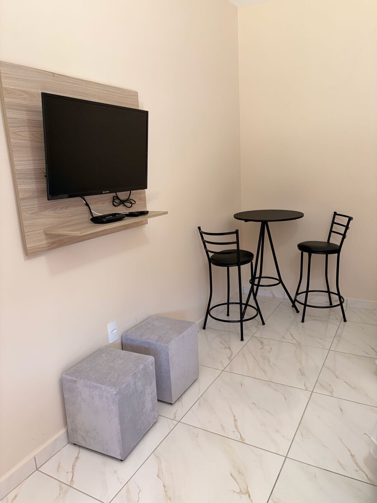 A small room with a flat-screen TV mounted on a wooden panel, two grey cushioned seating areas, and a round table with two chairs.