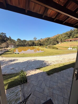 A view from inside a building looking out to a grassy area with a pond, a small shed, and a hill with trees in the background.