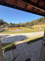 Vista desde el interior de un edificio a un área verde con un estanque, un cobertizo pequeño y una colina con árboles al fondo.