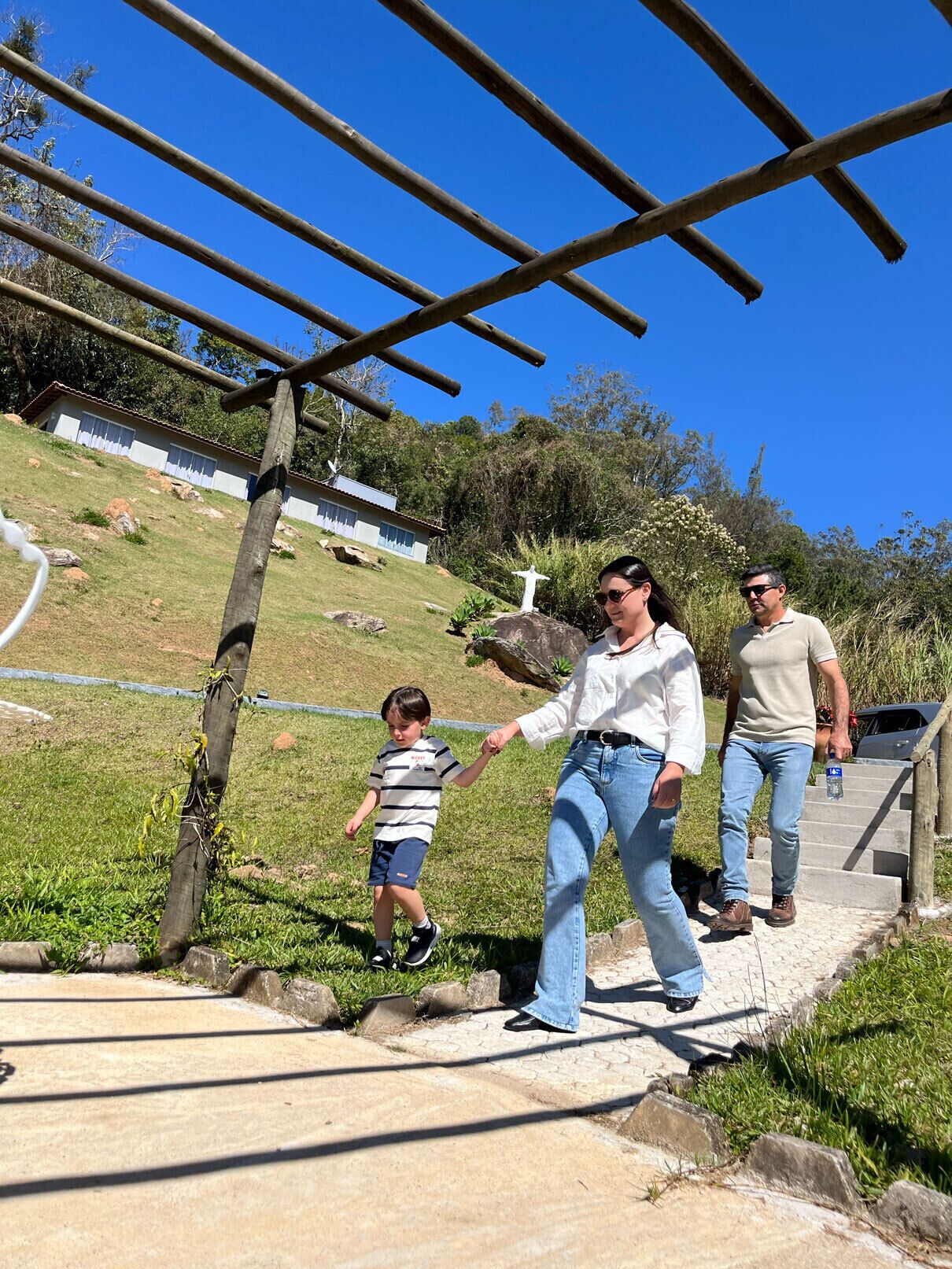 Um caminho com pérgula de madeira, uma área gramada e um edifício ao fundo.