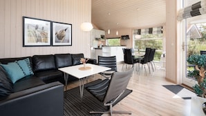 A modern living room with a black leather sofa, a white dining table, and black chairs. There is a kitchen in the background with white cabinets and a window. - Geraumiges Strandhaus in Grobenbrode (Grossenbrode)