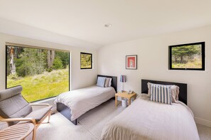 Habitación de hotel con dos camas, una silla, una mesita y grandes ventanales con vistas al bosque.