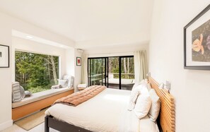 Ein modernes Schlafzimmer mit einem großen Bett, einer Sitzgelegenheit am Fenster und einem Balkon mit Blick auf Bäume.
