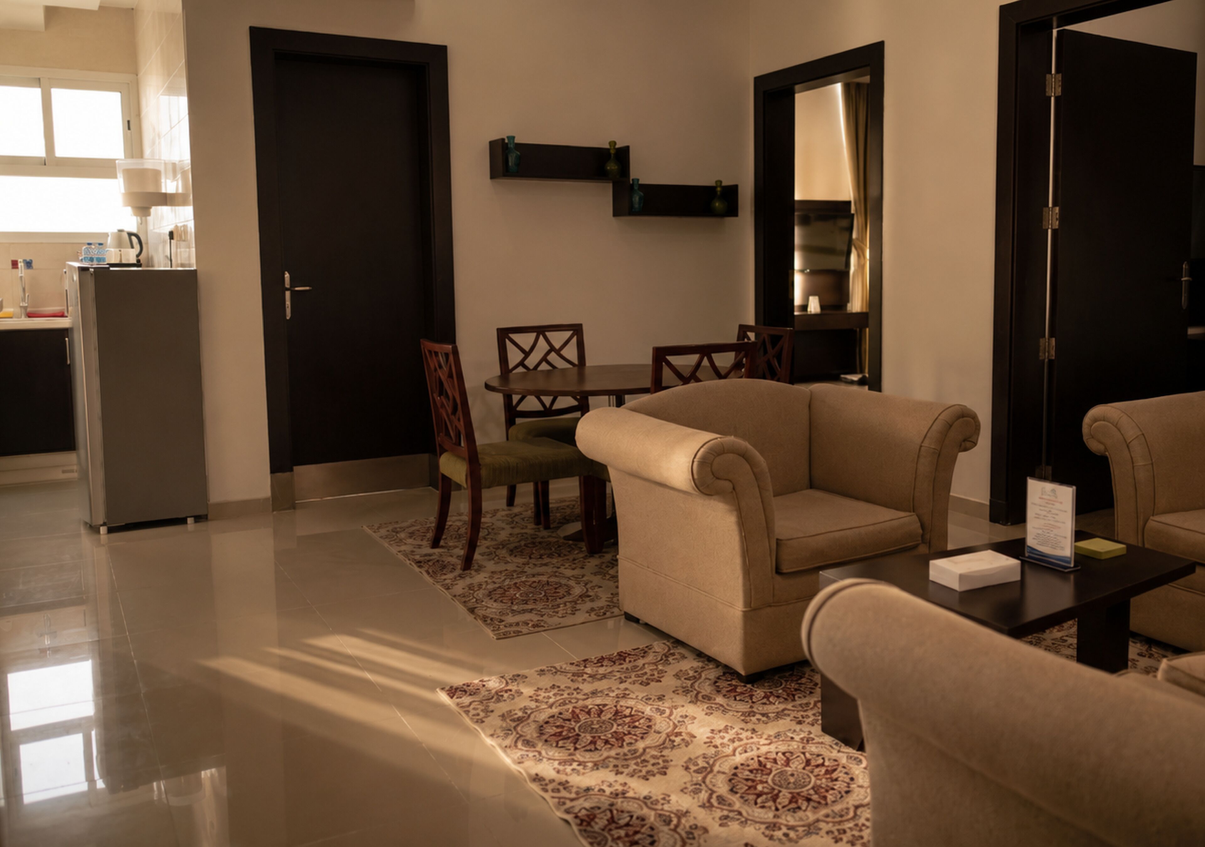 A modern living room with a sofa, chairs, a coffee table, and a rug. There is a kitchen area in the background with a refrigerator and cabinets.