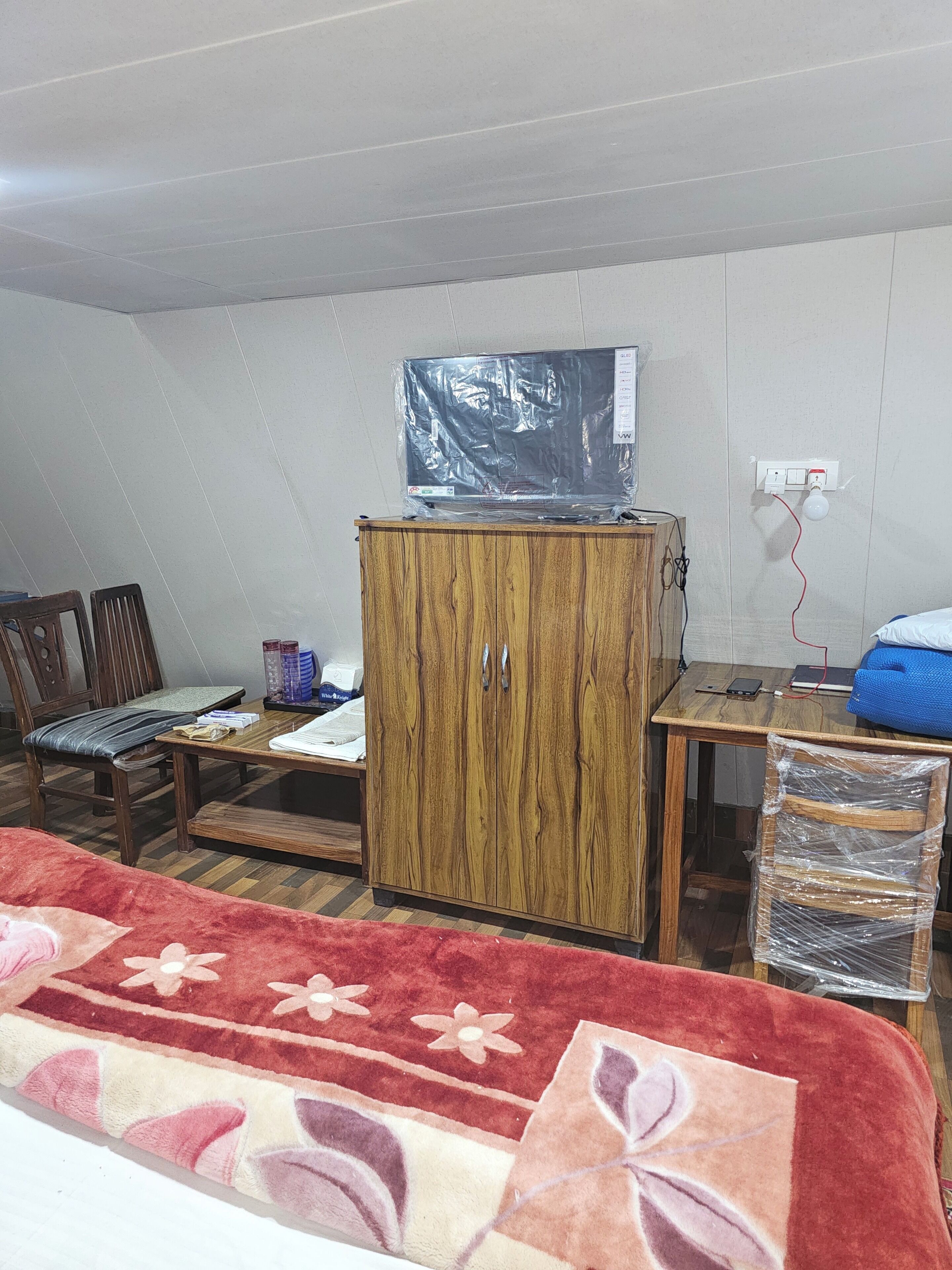 A room with a wooden cabinet, a television on top, a desk with a mobile phone, a chair, and a red patterned rug.