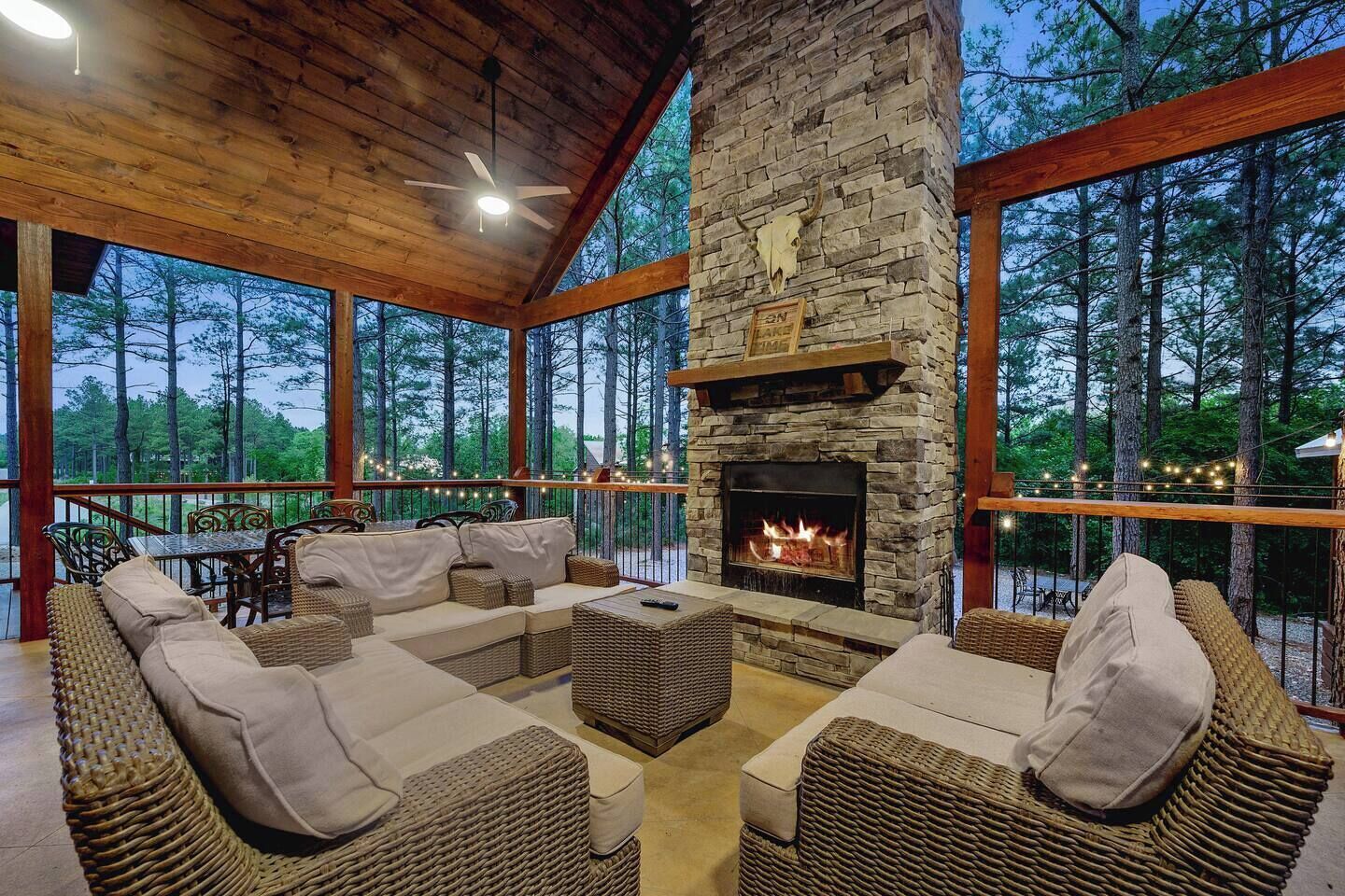 A covered outdoor seating area with a stone fireplace, wicker furniture, and a view of the surrounding forest.