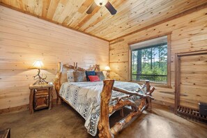 A wooden cabin bedroom with a bed centered on a log frame, two bedside lamps, a window with a view of trees, and a ceiling fan. - Dog-Friendly Lodge for 18 with 4 King Suites (Broken Bow)