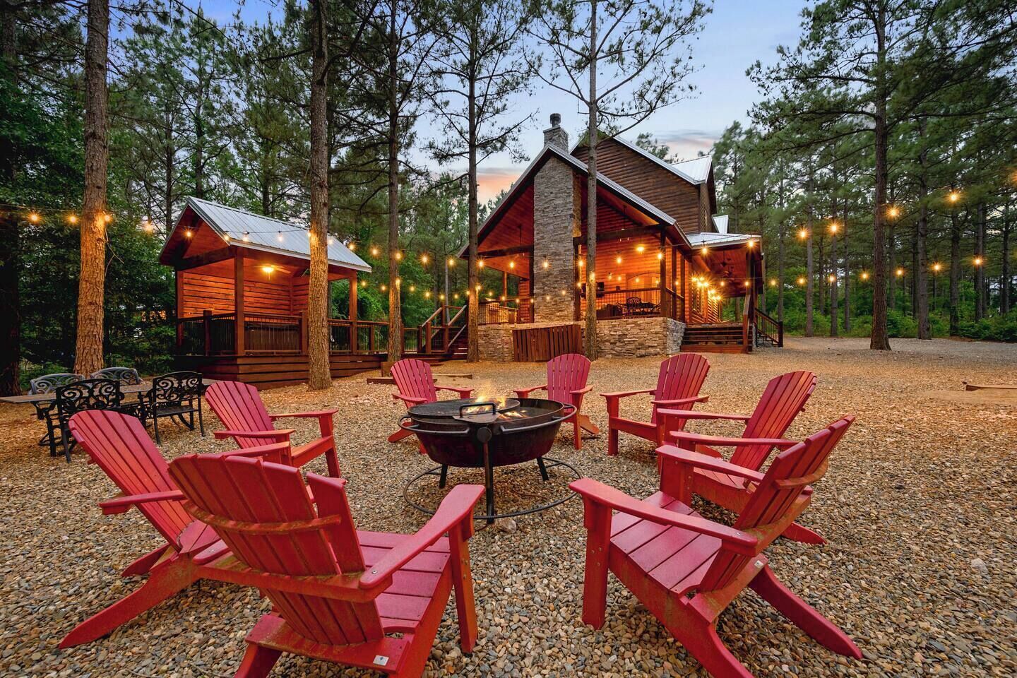 Une cabane dotée d’une cheminée en pierre, entourée de fauteuils Adirondack rouges, située dans une zone boisée et illuminée de guirlandes lumineuses.