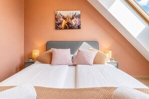 Un dormitorio con cama, almohadas, mesita de noche, lámpara y un cuadro colgado en la pared.