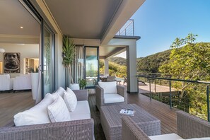 A modern outdoor seating area with wicker furniture, a glass door leading to a pool, and a view of a hilly landscape.