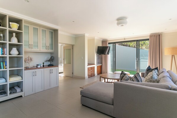 A modern living room with a grey sofa, a dining area with a table and chairs, and a kitchen with white cabinets and a built-in TV.