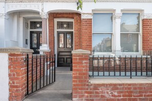 A traditional brick house with a black front door, a small porch, and a black metal fence. - Fulham Duplex Near Parsons Green Station (London)