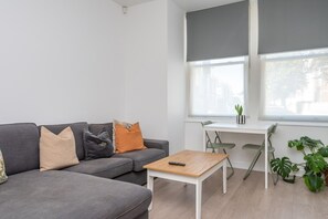 A modern living room with a grey sofa, a small wooden table, and a desk with a chair. There are two windows with blinds, a potted plant, and a view of buildings outside. - Fulham Duplex Near Parsons Green Station (London)