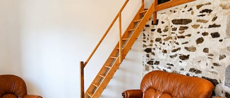 Una sala de estar con una pared de piedra, una escalera de madera, mobiliario de cuero y piso de baldosas.
