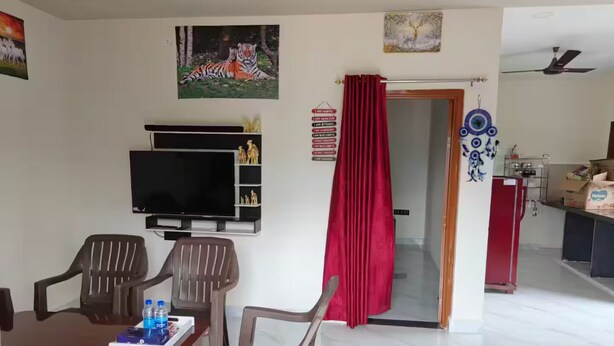 Ein Wohnzimmer mit Fernseher, einem Esstisch mit Stühlen, einem roten Vorhang und einer Küchenzeile im Hintergrund.