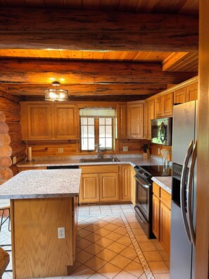A log cabin kitchen with wooden cabinets, a granite countertop, a stainless steel refrigerator, and a window with blinds. - Charming Log Cabin in the Heart of Amish Country (Millersburg)