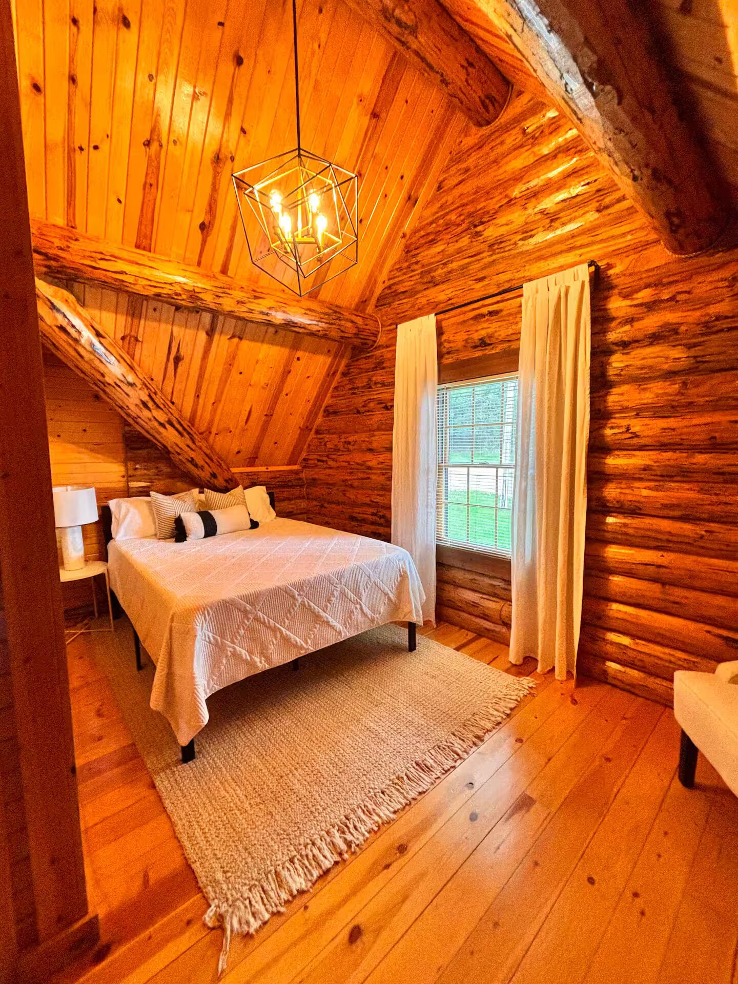 Une chambre à coucher dans un chalet en rondins, avec un lit, une table de chevet, une lampe, une fenêtre avec des rideaux et un luminaire suspendu.