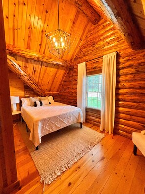 A log cabin bedroom with a bed, a nightstand, a lamp, a window with curtains, and a hanging light fixture. - Charming Log Cabin in the Heart of Amish Country (Millersburg)