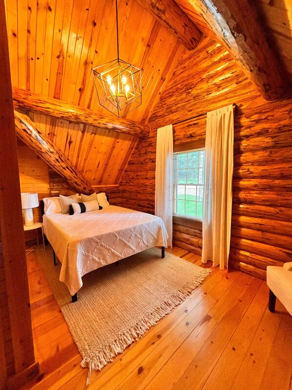 A log cabin bedroom with a bed, a nightstand, a lamp, a window with curtains, and a hanging light fixture.