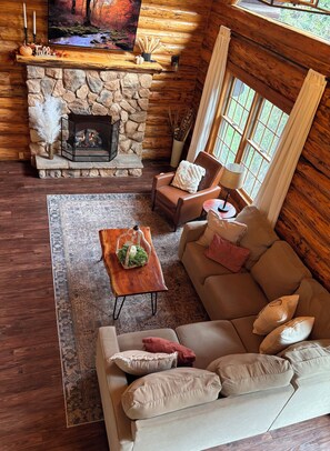 A cozy cabin interior with a stone fireplace, a leather sofa, a wooden coffee table, and a large window.