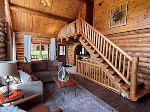 A log cabin interior with a staircase, a sofa, a coffee table, and a view of the outdoors. - Charming Log Cabin in the Heart of Amish Country (Millersburg)