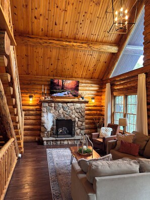 A log cabin interior with a stone fireplace, a television mounted above it, and a cozy seating area with a sofa and armchairs. - Charming Log Cabin in the Heart of Amish Country (Millersburg)