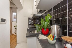 Eine Küche mit schwarzem Fliesenrückwand, einem Edelstahl-Wasserkocher, einer roten Vase mit Pflanze und einem Fenster mit Vorhängen.
