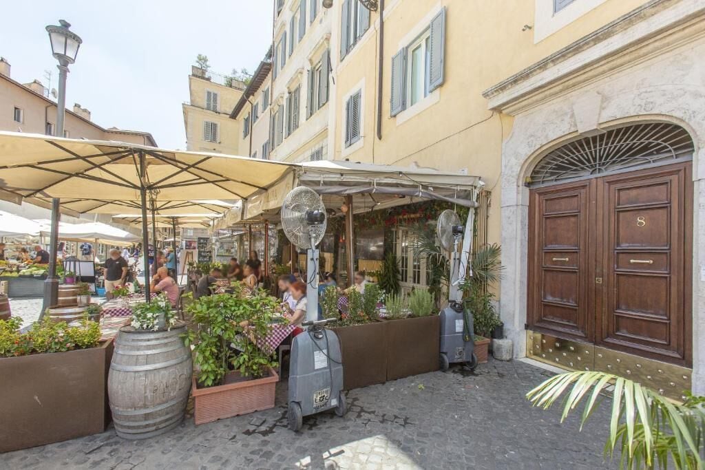 An outdoor café with tables and chairs, surrounded by potted plants and a large wooden barrel.