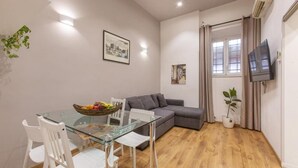 A compact living space with a glass dining table, white chairs, a grey sofa, and a flat-screen TV mounted on the wall. - SweetHouse Campo de Fiori (Roma)