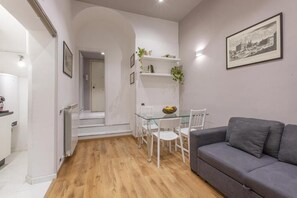 Ein modernes Wohnzimmer mit grauem Sofa, einem Glastisch mit Stühlen, einer weißen Treppe und einem gerahmten Bild an der Wand.