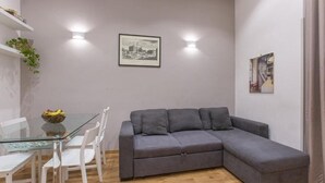 A modern living room with a glass dining table, white chairs, a grey sectional sofa, and two wall-mounted lights. - SweetHouse Campo de Fiori (Roma)