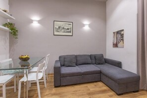 Ein modernes Wohnzimmer mit einem Glastisch, weißen Stühlen, einem grauen Ecksofa und zwei Wandleuchten.