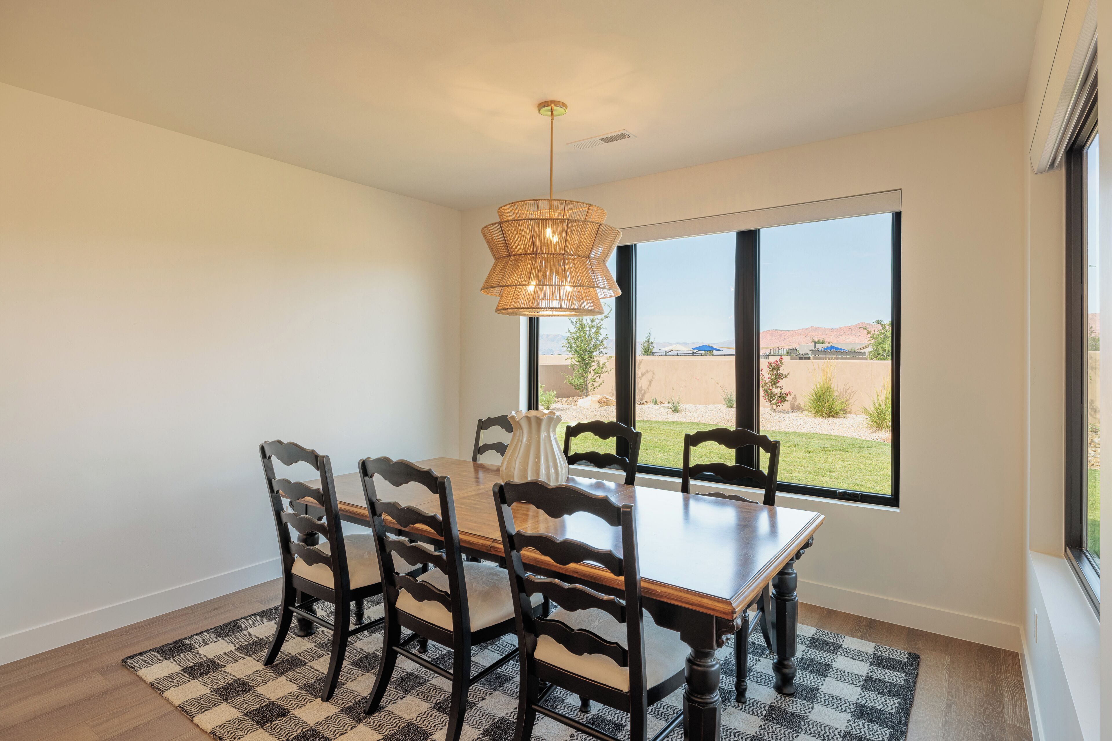 A dining room with a wooden table and chairs, a hanging light fixture, and a large window with a view of the outdoors.