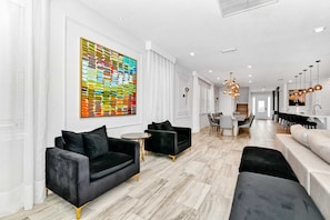 Ein modernes Wohnzimmer mit einem großen abstrakten Gemälde, einem Essbereich mit einer Kronleuchter und einer Küche mit Pendelleuchten.