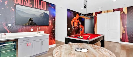 Una habitación con mesa de billar, televisor montado y una pared con un letrero que dice 'DOSE ALDO'.