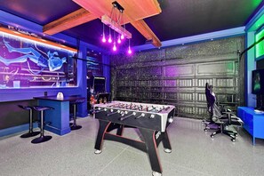 A room with a foosball table, a gaming chair, and a TV screen displaying a game. - UFC Champion José Aldo’s 9-BR Near to Disney (Kissimmee)