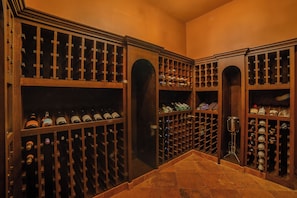 A wine cellar with multiple wooden racks for wine bottles, a metal wine rack, and a glass wine decanter on a pedestal. - Stunning 8BR Cabo Villa with Butler & Cooking Services (Cabo San Lucas)